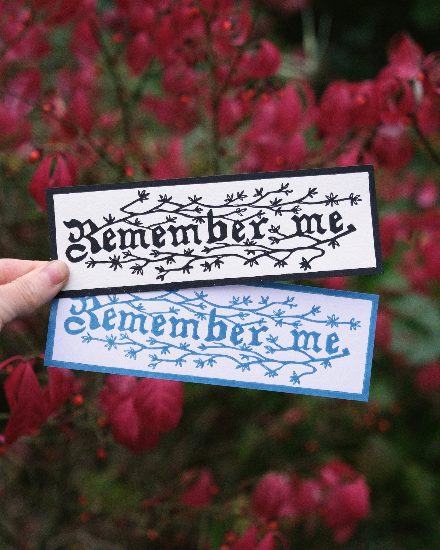 Two 'Remember me' bookmarks held against a blurred red leafy background