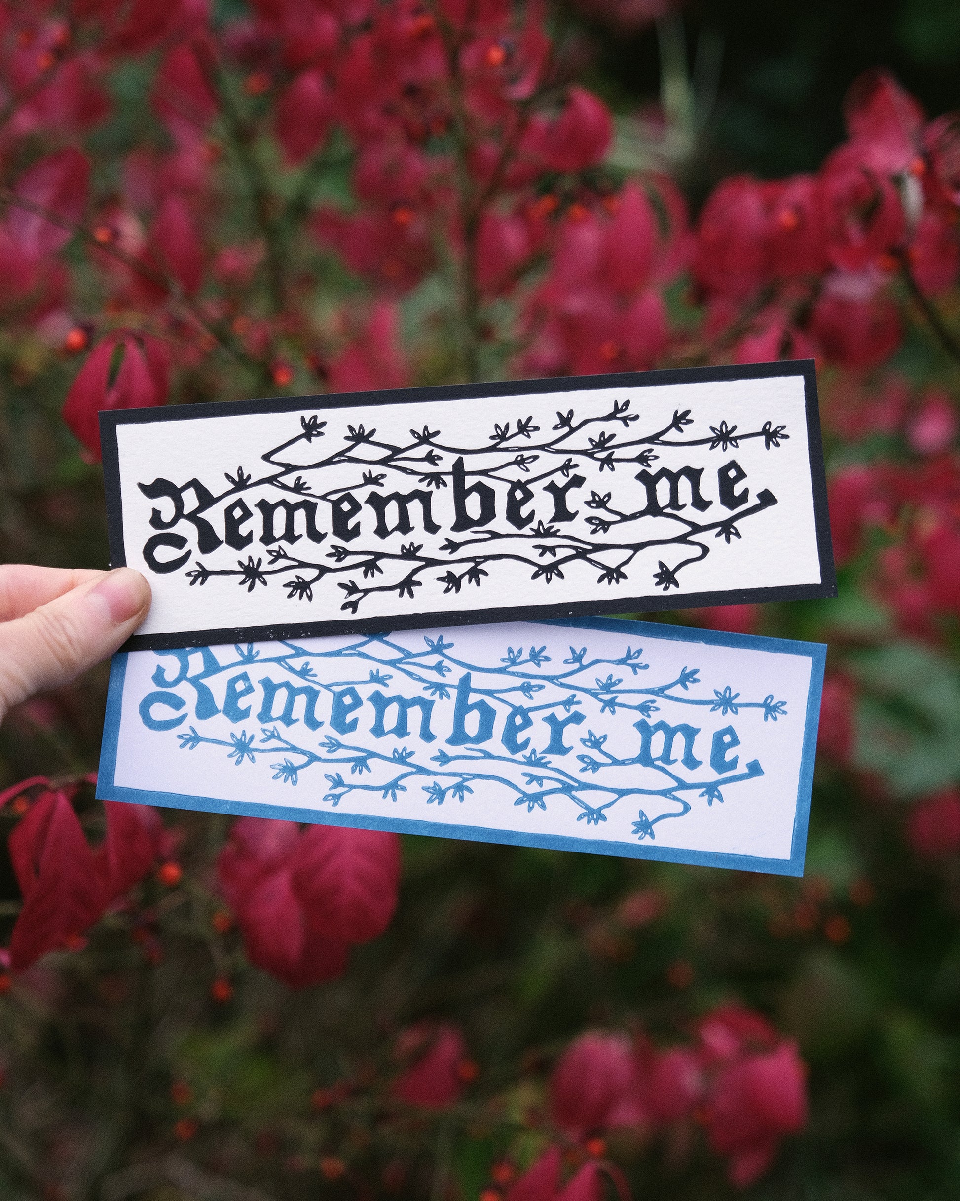 Two 'Remember me' bookmarks held against a blurred red leafy background